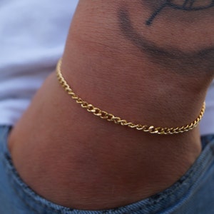 May include: A gold-colored chain bracelet with a curb link design. The bracelet is worn on a wrist, with a white shirt and blue jeans in the background. The bracelet is a simple, classic piece of jewelry.