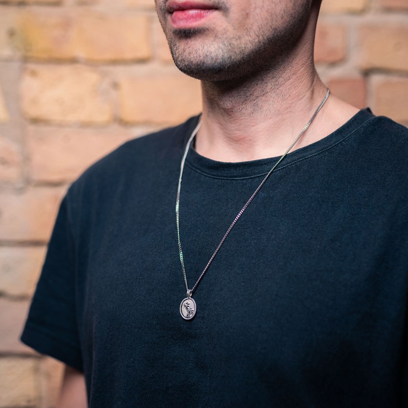 Pu&ograve; includere: Una collana d'argento con un piccolo ciondolo ovale con un motivo floreale. Il ciondolo &egrave; appeso a una catena delicata. La persona nell'immagine indossa una maglietta nera. Lo sfondo &egrave; un muro di mattoni.