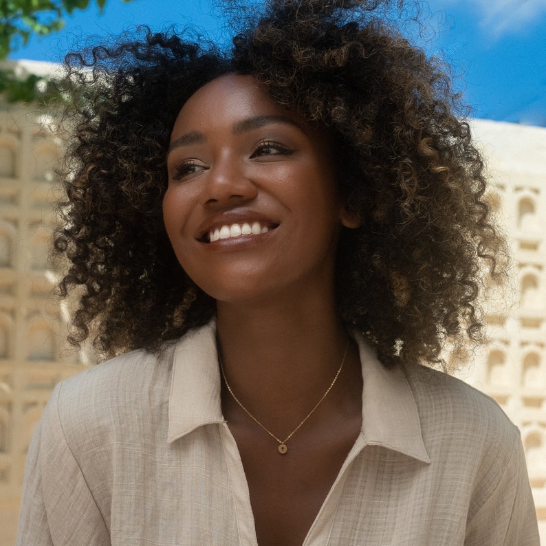 May include: A woman with curly brown hair is wearing a light brown shirt and a gold necklace with a small round pendant. She is smiling and looking up.