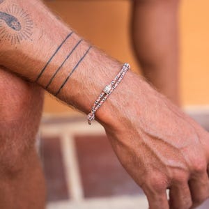 May include: A close-up of a person's wrist wearing a thin, braided bracelet with a silver bead. The bracelet is made of a light pink and white rope.