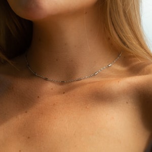 May include: A silver chain necklace with small rectangular links. The necklace is worn by a person with fair skin and blonde hair.