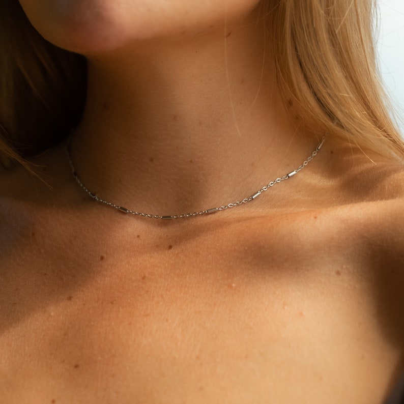 May include: A silver chain necklace with small rectangular links. The necklace is worn by a person with fair skin and blonde hair.