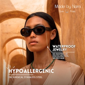 May include: A woman wearing black sunglasses with a gold chain attached to the frames. The sunglasses are resting on her nose. The chain is hanging down her chest. The woman is wearing a light brown shirt and a green beaded necklace. The text "WATERPROOF JEWELRY" is in a white box with a black outline. The text "NO RUSTING OR DISCOLORATION, NEVER FADE" is below the box. The text "HYPOALLERGENIC" is in a white box with a black outline. The text "316 SURGICAL STAINLESS STEEL" is below the box. The text "Made by Nami" is in the top right corner of the image. The text "Live Free" is below the text "Made by Nami".