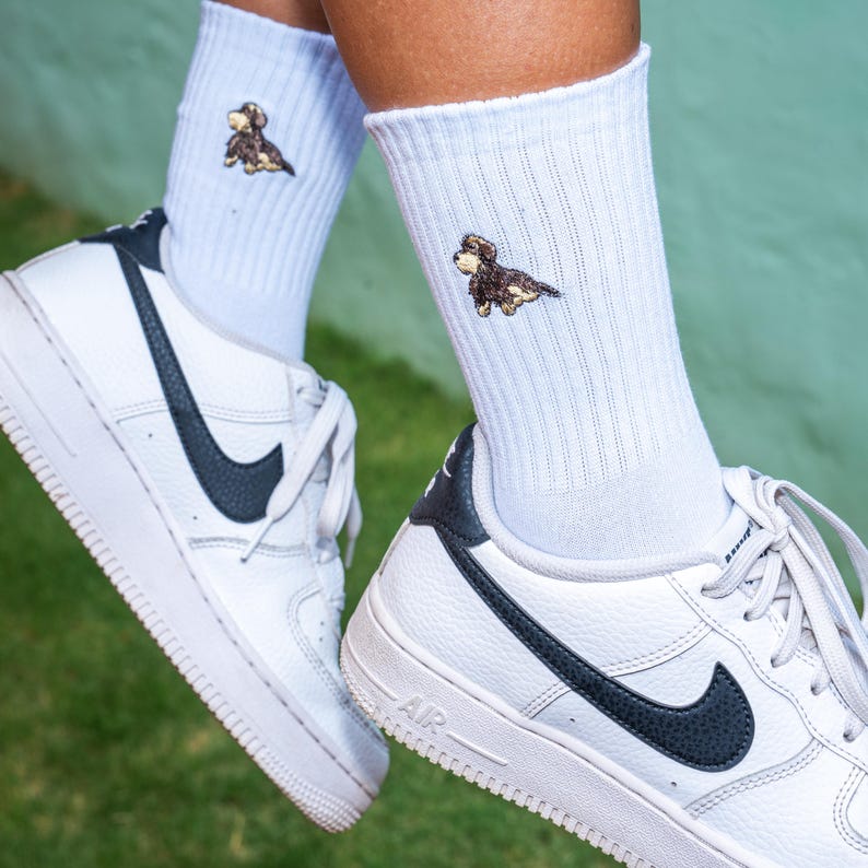 May include: White socks with a small embroidered dachshund on each sock. The socks are being worn with white trainers with black accents.