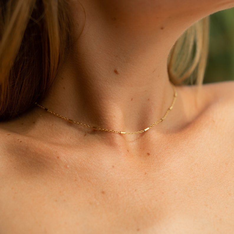 May include: A delicate gold chain necklace with small, rectangular links. The necklace is worn by a person with fair skin and blonde hair.