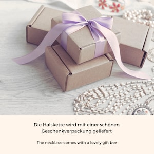 K&ouml;nnte beinhalten: Eine Geschenkbox mit einem lila Band und einer kleineren Geschenkbox im Inneren. Die Geschenkboxen stehen auf einer Holzoberfl&auml;che mit einer Perlenkette.