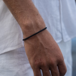 May include: A black rope bracelet is shown on a wrist. The bracelet is a simple, minimalist design, made of a tightly woven, dark material. The background is a white shirt.