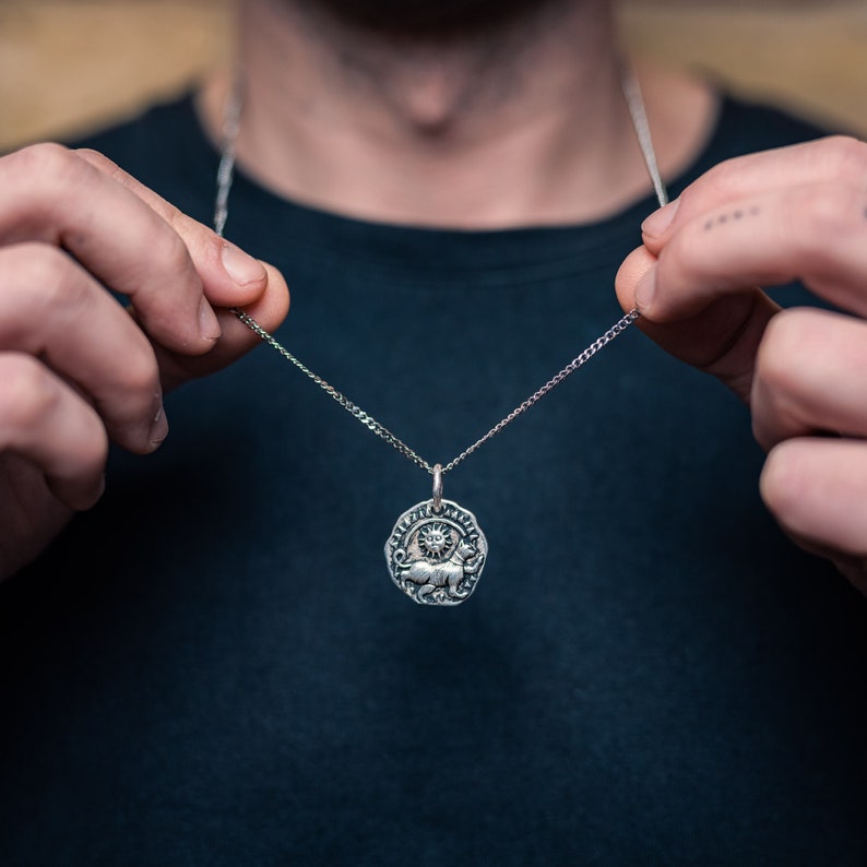 Pu&ograve; includere: Collana con pendente color argento con un pendente circolare con un disegno di sole e animale. Il pendente &egrave; appeso a una catena delicata. La collana &egrave; tenuta su uno sfondo scuro.