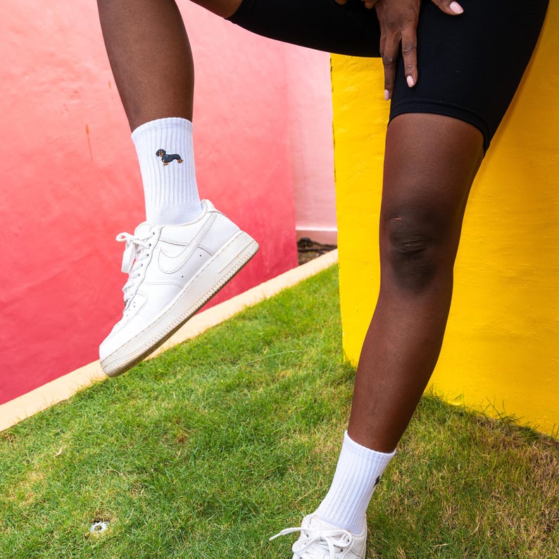 May include: A person wearing white socks with a dachshund graphic and white trainers against a pink and yellow background.