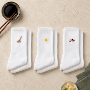 May include: Three pairs of white ribbed crew socks. The first pair features an embroidered sushi roll with chopsticks. The second pair displays an embroidered fried egg. The third pair shows an embroidered piece of nigiri sushi with a black seaweed strip.
