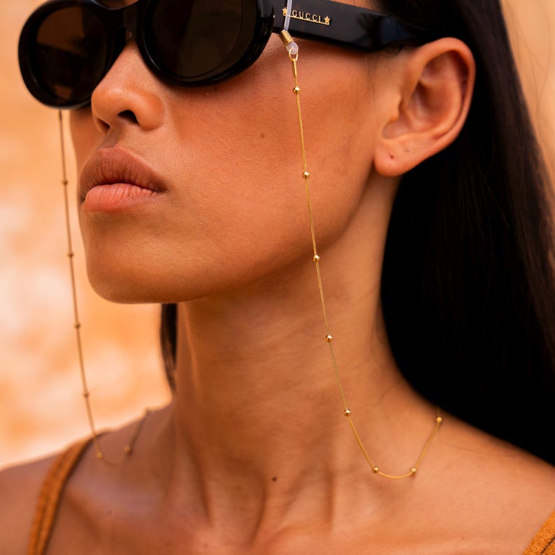 May include: A gold chain with small beads, designed to hold glasses. The chain is worn by a person with dark hair and black sunglasses.