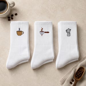 Könnte beinhalten: Drei weiße Crew-Socken mit gestickten Motiven rund um das Thema Kaffee. Die erste Socke zeigt eine braune Kaffeetasse, die zweite einen Siebträger mit Kaffeebohnen und die dritte eine silberne Espressokanne. Jede Socke hat ein geripptes Bündchen.