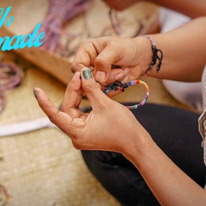 May include: A person's hands making a colorful friendship bracelet. The bracelet is made with colorful threads and is being tied together. The text "100% Handmade" is visible in the background.