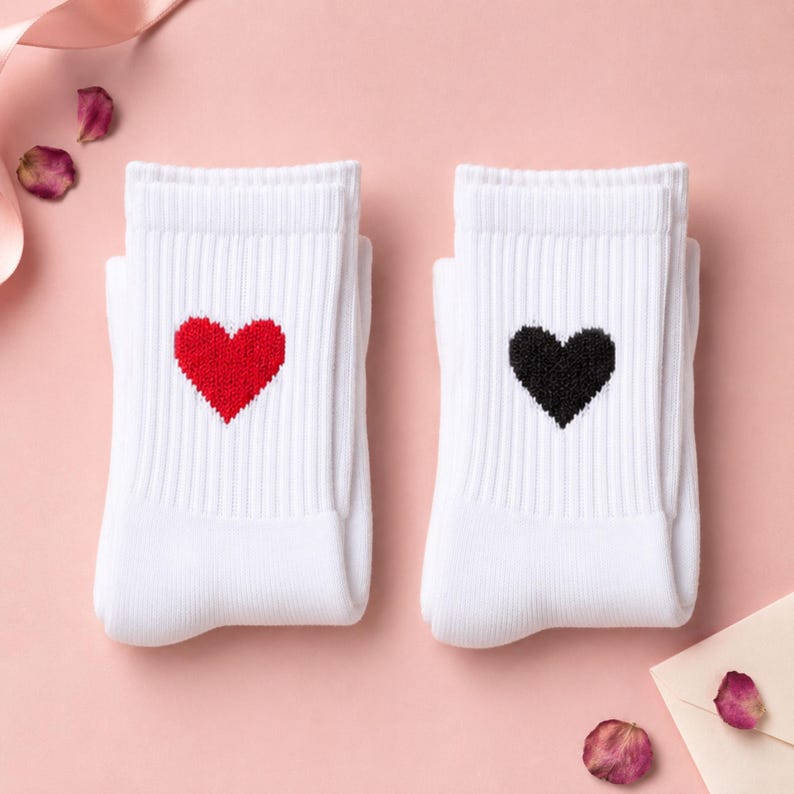 May include: A pair of white ribbed crew socks. One sock features a small, solid red heart embroidered on the upper section, while the other sock features a small, solid black heart embroidered in the same position on the white fabric.