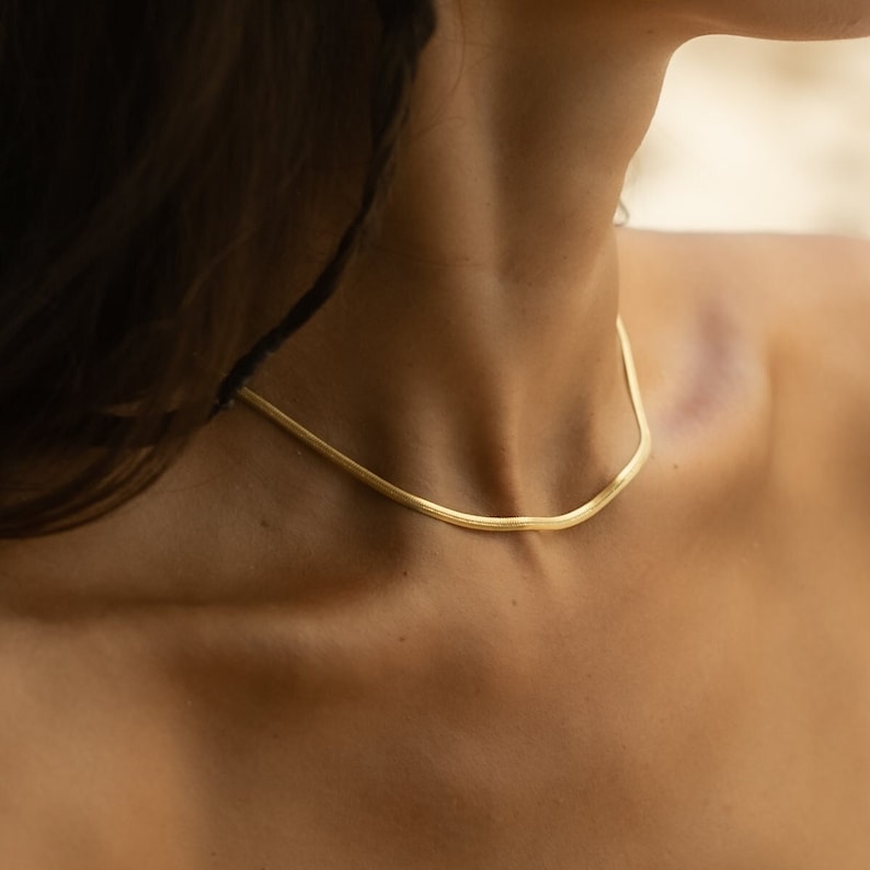 May include: A gold chain necklace with a simple, flat design. The chain is worn by a person with long brown hair.