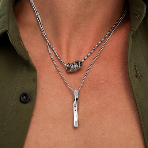 Może przedstawiać: Dwa srebrne naszyjniki. Górny naszyjnik ma trzy małe, połączone pierścienie. Dolny naszyjnik ma pionową, prostokątną zawieszkę. Naszyjniki są noszone na zielonej koszuli.