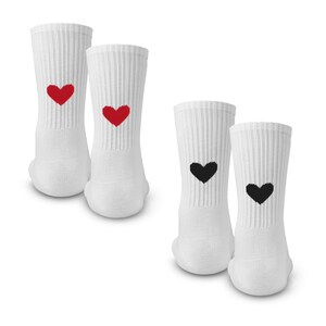 May include: Four white crew socks with ribbed texture. Two socks feature a small red heart graphic on the upper ankle area, while the other two socks feature a small black heart graphic on the same position. The socks are arranged in two pairs.