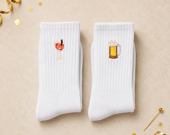 May include: A pair of white ribbed crew socks. The left sock features an embroidered orange cocktail in a stemmed glass, while the right sock displays an embroidered yellow beer mug with a white foamy head, both centred on the upper portion.