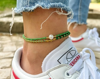 May include: A green and gold beaded anklet with a white pearl charm. The anklet is worn on a person's ankle.
