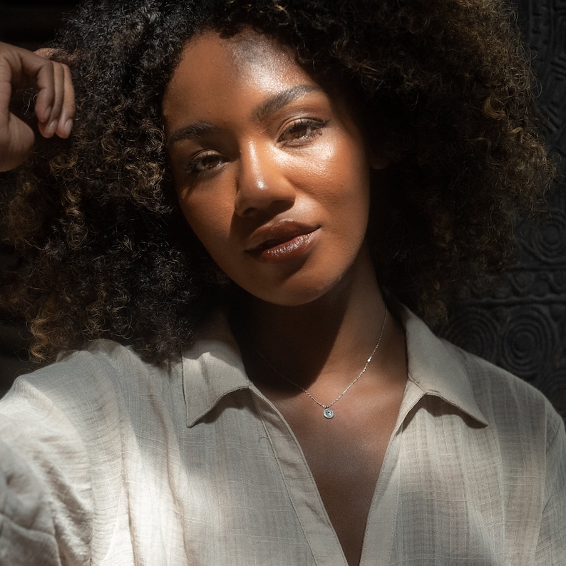 May include: A woman with curly brown hair wearing a light brown collared shirt and a silver necklace with a small round pendant.