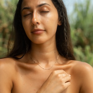 May include: A woman with long dark hair wearing a delicate gold chain necklace. The necklace is made of small, linked bars.