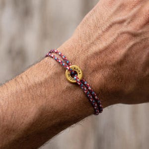 Könnte beinhalten: Ein Armband mit einer geflochtenen Kordel in Rot, Weiß und Blau, die durch einen kleinen, runden, goldfarbenen Metallanhänger gezogen ist. Der Anhänger ist mit dem Namen Anna und dem Datum 04.12.09 in einer klaren, dunklen Schrift graviert.