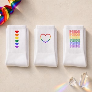 Pode incluir: Três meias brancas de cano médio com designs coloridos. A primeira meia tem uma coluna vertical de seis pequenos corações com as cores do arco-íris. A segunda exibe o contorno de um coração arco-íris. A terceira apresenta a palavra PRIDE em seis linhas coloridas.
