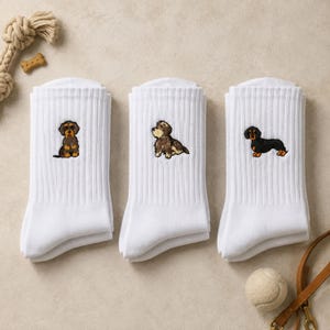 Könnte beinhalten: Drei Paar weiße, gerippte Socken, jeweils mit einer kleinen Stickerei eines Dackels. Die erste Socke zeigt einen braun-schwarzen Rauhaardackel, die zweite einen braun-weißen Hund und die dritte einen schwarz-lohfarbenen Dackel.