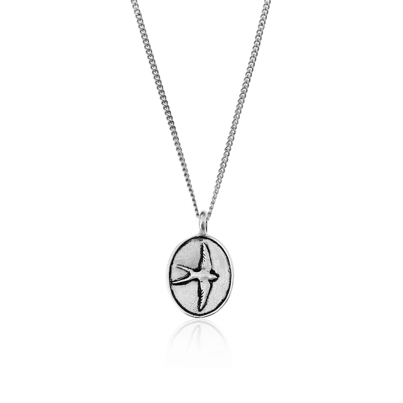 May include: A silver chain necklace with a pendant featuring a stylized image of a swallow in flight. The pendant is oval shaped and has a textured surface.