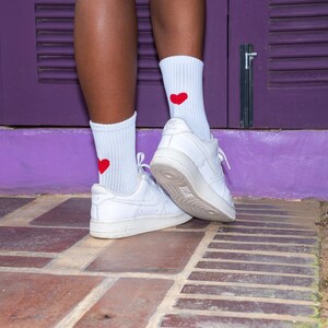 May include: A pair of white ribbed crew socks featuring a small, solid red heart embroidered on the outer side of each sock. The socks are worn with white leather low-top sneakers that have a thick rubber sole and a lace-up closure system.