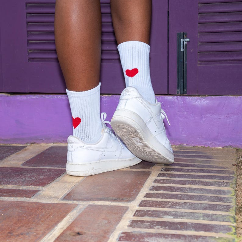 May include: A pair of white ribbed crew socks featuring a small, solid red heart embroidered on the outer side of each sock. The socks are worn with white leather low-top sneakers that have a thick rubber sole and a lace-up closure system.