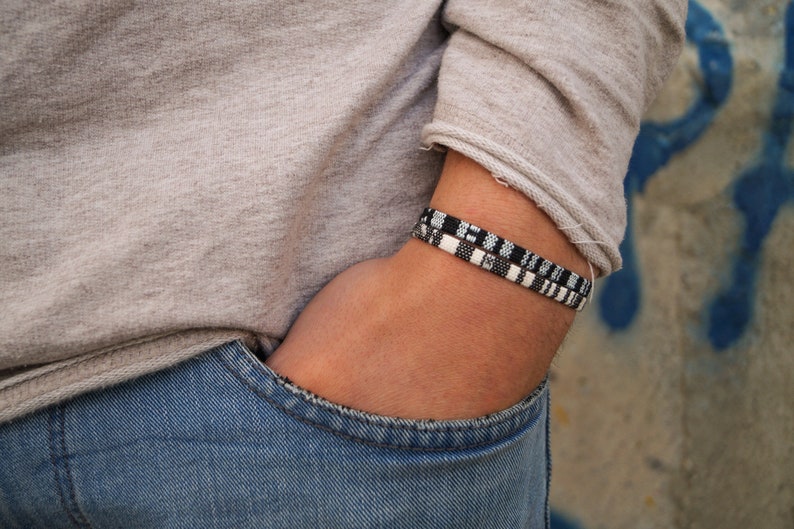 May include: A person wearing a gray shirt and blue jeans with a black and white woven bracelet on their wrist.