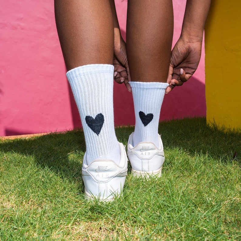 May include: A pair of white ribbed crew socks featuring a small black heart graphic on the back of each ankle, paired with white low-top athletic sneakers. The socks are made of a textured knit fabric and reach mid-calf, showcasing a simple design.