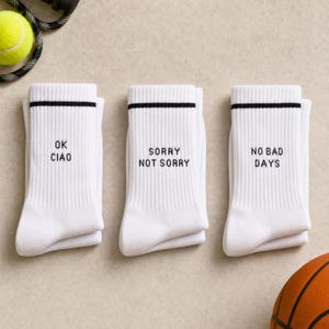 May include: Three pairs of white ribbed crew socks featuring black text and a single black stripe near the top. The first pair reads OK CIAO, the second pair reads SORRY NOT SORRY, and the third pair reads NO BAD DAYS in a simple black font.