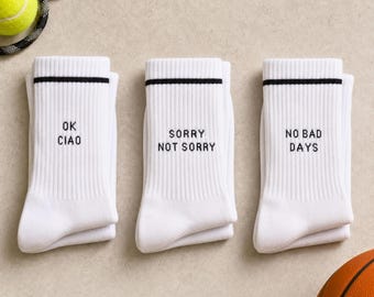 May include: Three pairs of white ribbed crew socks featuring black text and a single black stripe near the top. The first pair reads OK CIAO, the second pair reads SORRY NOT SORRY, and the third pair reads NO BAD DAYS in a simple black font.
