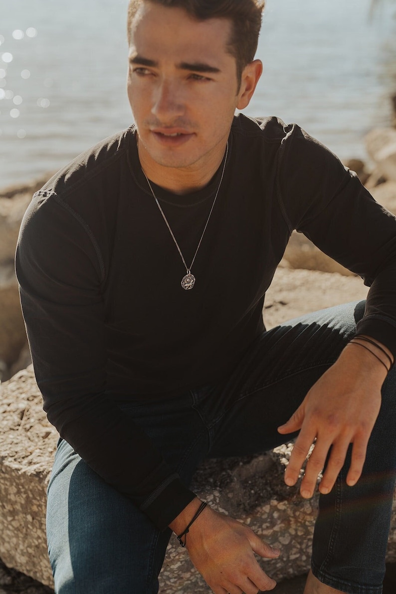 Pu&ograve; includere: Una collana con pendente in argento con un design circolare &egrave; appesa a una catena. La persona nell'immagine indossa una maglietta nera a maniche lunghe e jeans blu. Lo sfondo &egrave; sfocato, con uno specchio d'acqua e rocce.