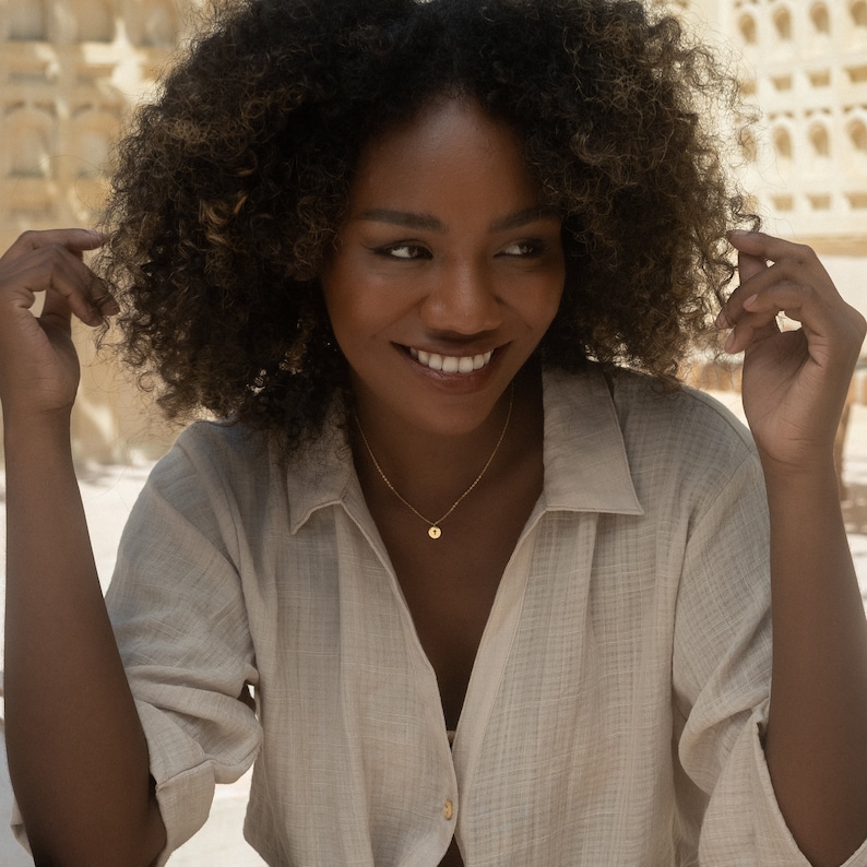 May include: A woman with curly brown hair wearing a light beige shirt with a gold necklace and a small round pendant.