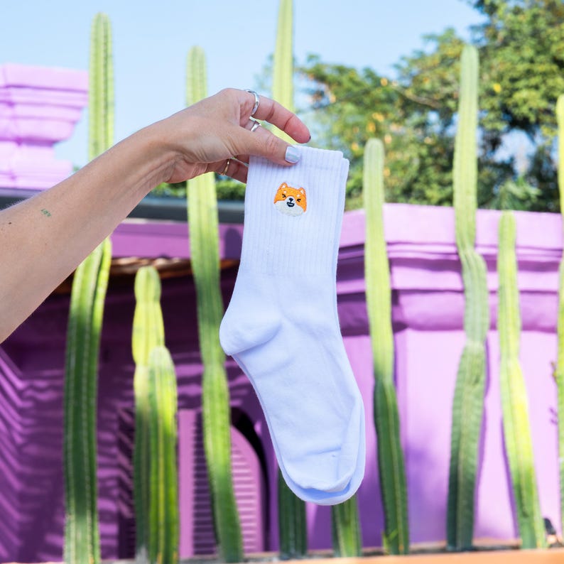 K&ouml;nnte beinhalten: Wei&szlig;e Socken mit einem niedlichen gestickten Shiba Inu-Gesicht auf der Vorderseite. Die Socken werden vor einem lila und gr&uuml;nen Hintergrund gehalten.