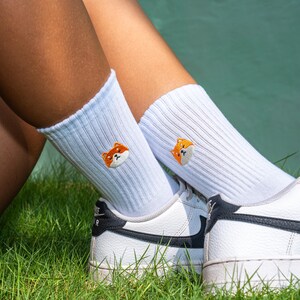 K&ouml;nnte beinhalten: Wei&szlig;e Socken mit gestickten orangefarbenen Shiba Inu-Gesichtern. Die Socken werden mit wei&szlig;en Sneakers mit schwarzen Akzenten getragen.