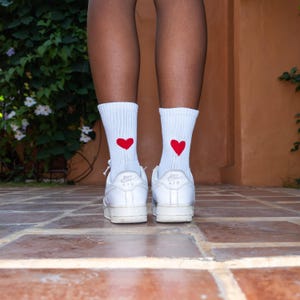 May include: A pair of white ribbed crew socks featuring a small red heart graphic on the back of each ankle, paired with white leather low-top sneakers that have the word AIR embossed on the heel and sole.