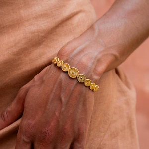 Puede incluir: Un brazalete dorado con una serie de discos redondos texturizados. El brazalete se lleva en la muñeca.