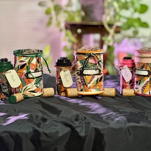 May include: Four colourful, decorated metal cans with lids, each with a tag tied to the handle. The cans are decorated with a tropical leaf pattern in shades of green, orange, pink, and white. The tags are blank, and the cans are sitting on a black surface.