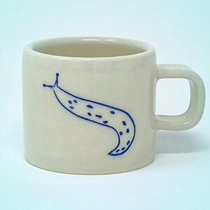 Könnte beinhalten: Cremefarbene Keramik-Tasse mit einer blauen Illustration einer Schnecke. Die Tasse hat einen abgerundeten Körper und einen quadratischen Griff. Das Schnecken-Design hat eine einfache Kontur mit gepunkteten Details.