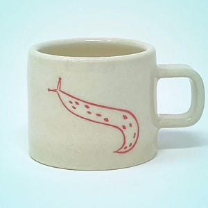 May include: Cream-colored ceramic mug with a square handle. A red, stylized illustration of a slug is on the side. The mug has a cylindrical shape and a smooth, glossy finish. A unique and whimsical design for coffee or tea.