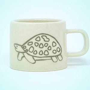 May include: A white ceramic mug with a hand-drawn outline of a turtle with a patterned shell. The turtle is facing right and has a simple, whimsical design.