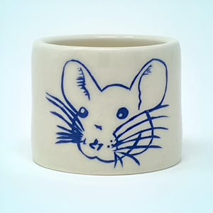 May include: A cylindrical ceramic cup with a cream-colored base. The cup features a blue illustration of a mouse's face. The mouse has large ears, whiskers, and small eyes. The cup is suitable for holding beverages or small items.