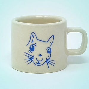 May include: A cream-colored ceramic mug with a blue squirrel face design. The mug has a square handle and a cylindrical shape. The squirrel face is hand-drawn with blue lines, featuring large eyes and whiskers. The mug is suitable for hot or cold beverages.