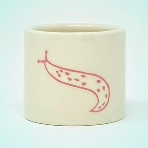 May include: A cylindrical, off-white ceramic mug featuring a pink illustration of a slug. The slug design has a dotted pattern and is outlined in pink. The mug has a smooth, matte finish and a simple, minimalist design.