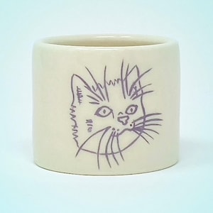 May include: A cylindrical, off-white ceramic cup with a hand-drawn illustration of a cat's face in purple ink. The cat's features include large eyes, whiskers, and a slightly open mouth. The cup is smooth and has a simple, rounded shape.