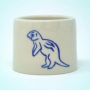 Puede incluir: Taza cilíndrica de cerámica color crema. Un dibujo azul de una foca está en el lateral. La taza tiene un acabado liso y brillante y un diseño sencillo y minimalista.